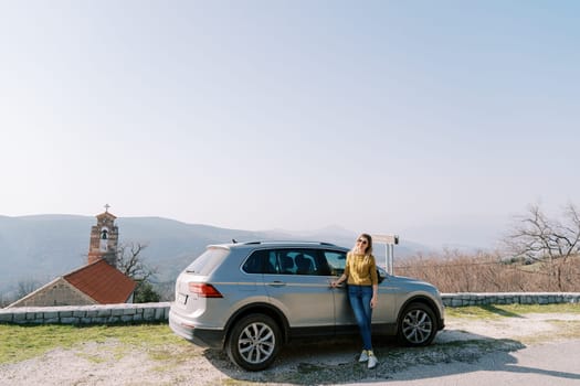 Smiling woman stands near the car on the observation deck next to the church in the mountains. High quality photo