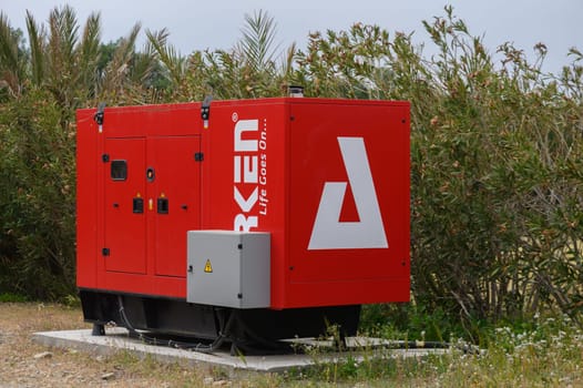 Gaziveren Cyprus -17.03.2025 A vibrant red generator is placed on a concrete base, tucked among lush green plants. It stands ready to provide power in a serene outdoor environment under gray clouds.
