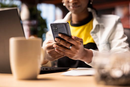 Woman reading messages on phone in living room, having discussion with mates, close up shot. African american person in apartment office reading SMS on cellphone device, chatting with friends