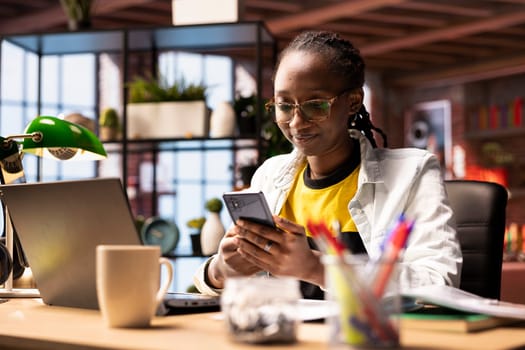 Smiling woman in home office using smartphone to compose social media posts, producing content. African american person monitoring social media engagement using mobile phone