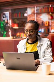 Freelancer in home office using laptop device to work on job tasks. African american remote employee using notebook to do customers relations job online, answering inquiries