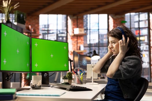 Female employee uses green screen mockup and headset to finish tasks in a creative agency office setup, high tech work environment. Modern workstation enhances productivity and business efficiency.