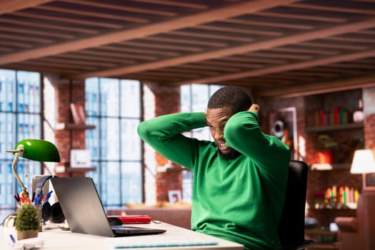 Teleworker in home office feeling hurting pain on the back of the head while working on laptop. Jaded remote employee suffering neck injury while solving tasks on notebook