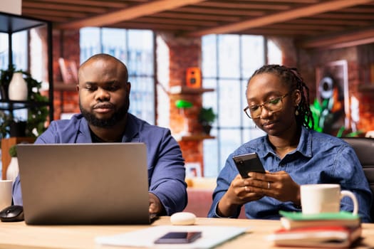 Black couple working remotely on daily freelancing tasks at a desk, young man studying reports on laptop and woman networking with clients on smartphone. Teamwork for efficiency.