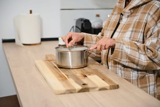 A woman in a plaid hoodie stirs a saucepan on a wooden board in a modern kitchen. The scene exudes warmth and coziness with clean design and home-cooking ambiance.
