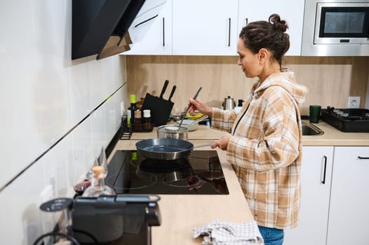 A young woman prepares food in a modern kitchen with an induction stove. She appears focused while holding utensils and cooking on the counter. A cozy home kitchen setting is visible.