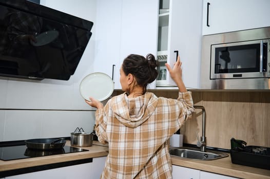 A young woman reaches to organize a cabinet while holding a plate in a modern kitchen. The space reflects a combination of functionality and comfort, embodying everyday household activities.
