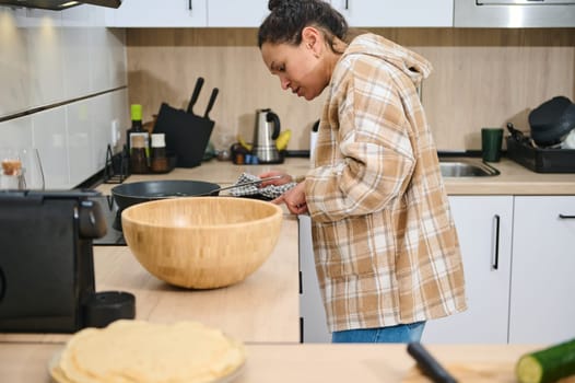 A young woman prepares food in her home kitchen, focusing on healthy and homemade meals. Kitchen items and ingredients are visible, portraying a modern and cozy cooking environment.