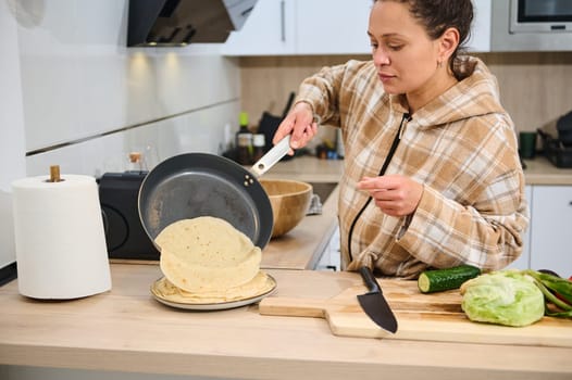 A young woman cooks fresh tortillas while preparing raw ingredients for a salad in her kitchen. The image emphasizes homemade cooking, healthful living, and domestic activity.