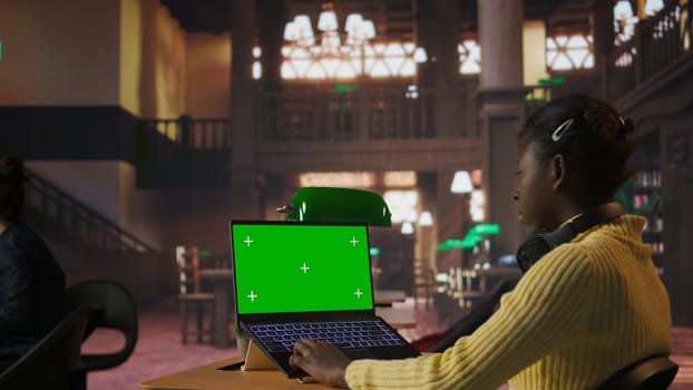 Black scholar reading information via internet next to chroma key in a library, focusing on theoretical studies and completing her class assignments. Taking notes for educational work. Camera A.