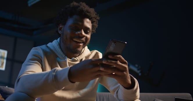 Relaxed black freelancer chatting with friends on his smartphone via texts, sitting on the couch. Young adult showcasing laughter and leisure, staying entertained while working remotely. Camera B.