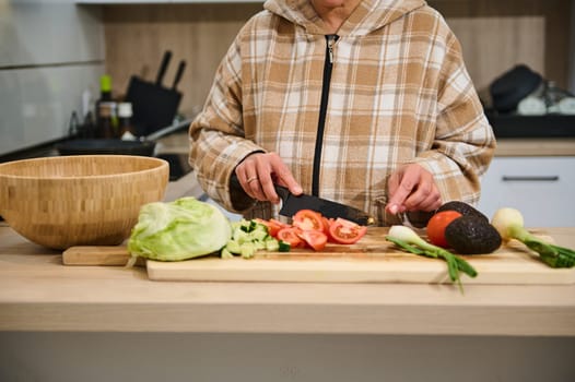 A person is slicing vegetables, including tomatoes and lettuce, on a wooden board for a salad. Various fresh ingredients are visible, showcasing a healthy cooking atmosphere and lifestyle.