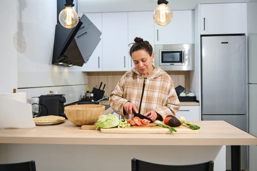 A woman slices fresh vegetables in her home kitchen, creating a vibrant and healthy salad. The modern kitchen setup exudes a sense of warmth, healthiness, and culinary creativity.