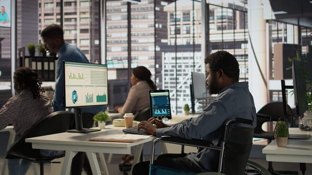 African american male worker in wheelchair trying to solve daily tasks at the office, examining organizational data and working on a forecasting plan. Man with impairment ensures analysis. Camera B.