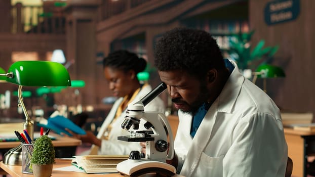 Male student researching viruses during a microscope analysis and academic reading in a classic library. Med school pupil reflects dedication to biology and exam preparations. Camera B.
