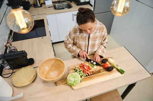A young woman slicing vegetables for a salad in a bright, modern kitchen. The setting showcases a cozy and fresh cooking atmosphere, emphasizing healthy eating and wholesome meal preparation.