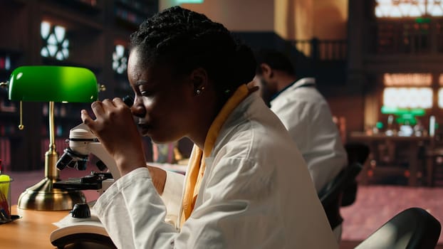 Black university student examining cells and organisms under microscope lenses, researching and studying for medical science exams. Reading about biotechnology in the library. Camera A.