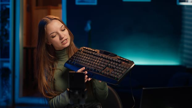 Gen Z girl presenting RGB lit mouse and keyboard while doing review in personal studio to see if price is justified. Young influencer showcasing neon lit computer peripheral features to fans, camera A