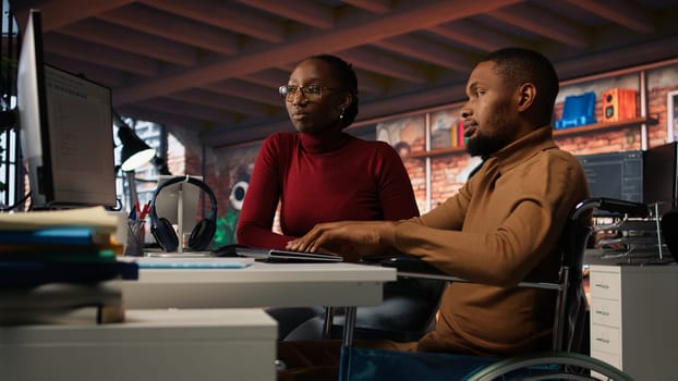 Manager coming to inclusive startup office employee in wheelchair desk to offer help. Thoughtful supervisor checking up on software development agency worker with disability, camera A
