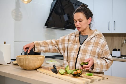 A woman in a checkered jacket prepares an avocado salad in her modern kitchen, showcasing a cozy and healthy lifestyle. The scene emphasizes fresh, nutritious ingredients and home cooking.