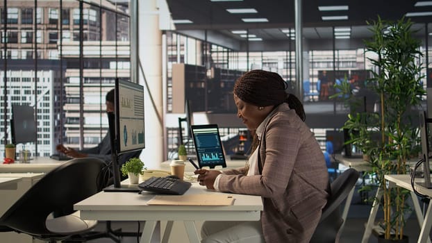 Black business employee browses social media webpages on her phone, taking a short break to answer a few texts at the office. Female worker does online communication via texting. Camera B.