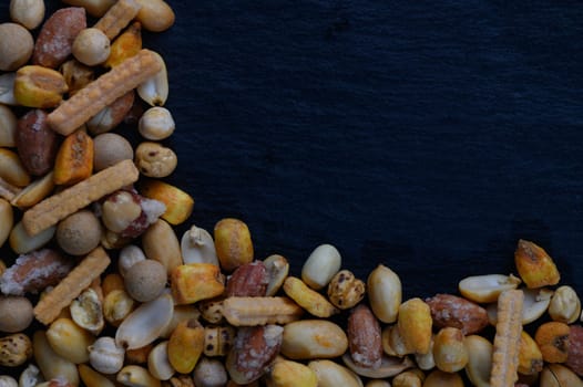 A vibrant mix of nuts and crispy snacks is artfully scattered along the edges of a dark slate surface, inviting sharing and enjoyment during lively conversations.