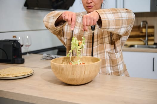 Young woman tosses fresh vegetables in a wooden bowl in a cozy kitchen for a healthy meal.