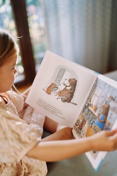 Little girl is sitting on the bed reading a fairy tale about the beaver workshop with pictures. Side view. Cropped. High quality photo