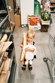 Little girl with a toy hare and a big baguette in a package stands in a supermarket. High quality photo