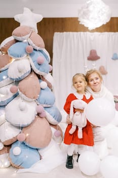Little girl with a balloon stands near an actress with small deer antlers near a toy Christmas tree on stage. High quality photo