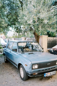 Vintage car Zastava Skala stands under a green tree near a stone fence. High quality photo