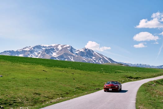 Red convertible drives along a highway in a valley with snow-capped mountains in the background. High quality photo