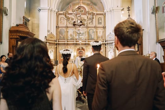 Guests stand in the church behind bride and groom in wreaths in front of the priest during the wedding ceremony. High quality photo