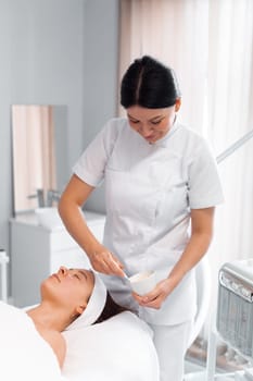 Dermatologist in uniform preparing face mask for female client lying on massage table at salon. Relaxed young woman undergoing facial treatment in beauty spa