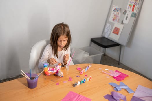 School-aged girl with blonde hair sitting at table at home, painting plaster figurines with glue and colorful gouache paints, engaging in creative arts and crafts for child development