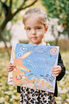 Little girl with a colorful book in her hands stands in the garden. Title: Stormy weather. Debi Gliori. High quality photo