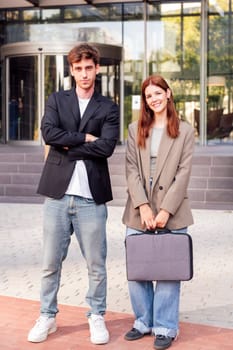 business man and woman standing outside of an office building smiling happy while looking at camera, they are wearing casual clothes and she has a briefcase