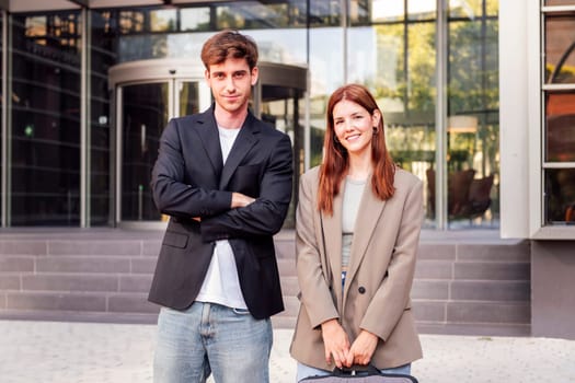 business man and woman standing outside of an office building smiling happy while looking at camera, they are wearing casual clothes and she has a briefcase
