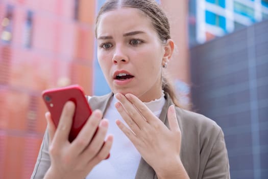 Caucasian woman looking at her smartphone with a shocked and surprised expression. Unexpected news, social media reaction, and technology concept in an urban environment with modern buildings.
