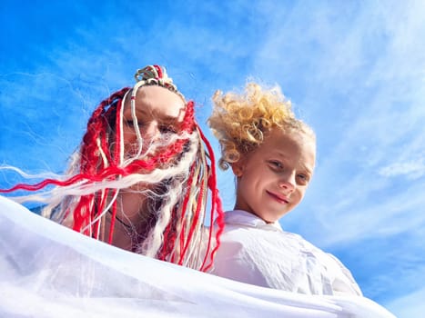 Two individuals with vibrant hairstyles smile and playfully hold a white fabric against a backdrop of blue sky.