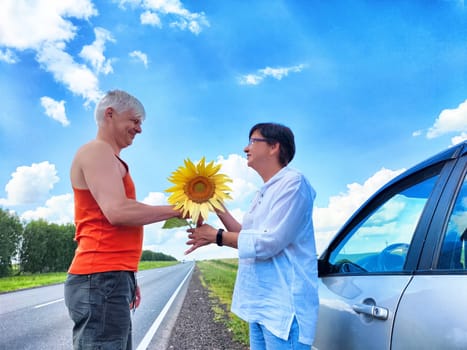 A happy couple exchanges a bright sunflower by the roadside, surrounded by lush greenery and a clear blue sky, enjoying their travel adventure together.