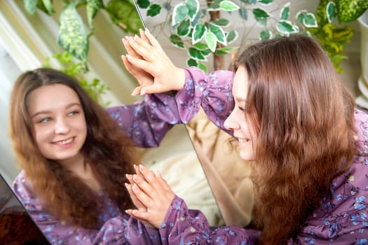 A girl with curly hair enjoys a moment of reflection, smiling and engaging with her mirror image in a lively indoor garden.