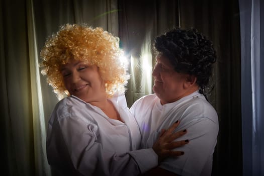 A middle-aged couple poses in an apartment, showcasing their love and fun while wearing vintage wigs during a theater-inspired photoshoot.