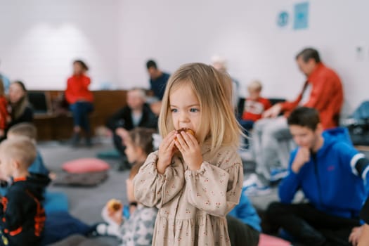 Little girl stands in the kindergarten hall and eats a bun. High quality photo