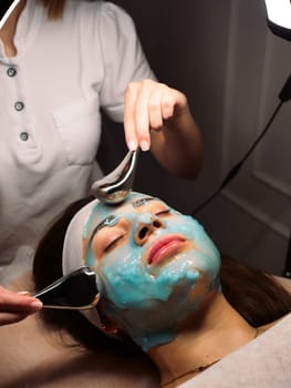 Cosmetologist does cryo massage with globules on a blue hydrogel alginate mask in the beauty salon, vertically, close-up. Woman Facial Skin and Body Care