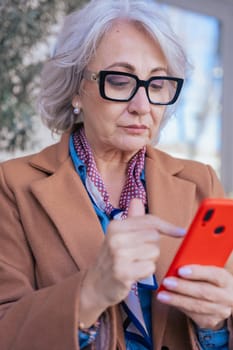 Confident senior caucasian woman with gray hair and glasses using a smartphone outdoors.Modern lifestyle, digital technology,and sophistication in an urban setting with elegant fashion and accessories