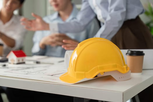 A yellow hard hat symbolizes safety in a collaborative setting, while team members discuss architectural designs and project details over blueprints and models.