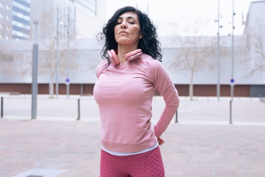 Caucasian adult woman with curly black hair stretching her arms outdoors in an urban setting.she is wearing a pink sweatshirt and exercising for a healthy lifestyle, fitness, and wellness in the city