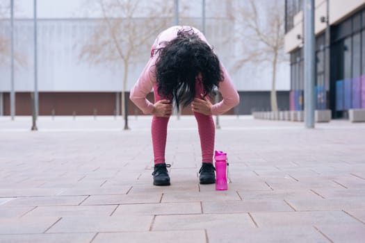 Caucasian adult woman with curly black hair stretching her arms outdoors in an urban setting.she is wearing a pink sweatshirt and exercising for a healthy lifestyle, fitness, and wellness in the city