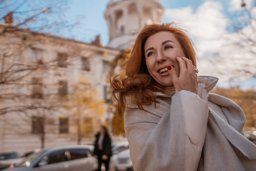 Redhead woman in a beige coat smiles, hand on face, outdoors. Old European architecture and parked cars blur in the background. Autumnal setting.